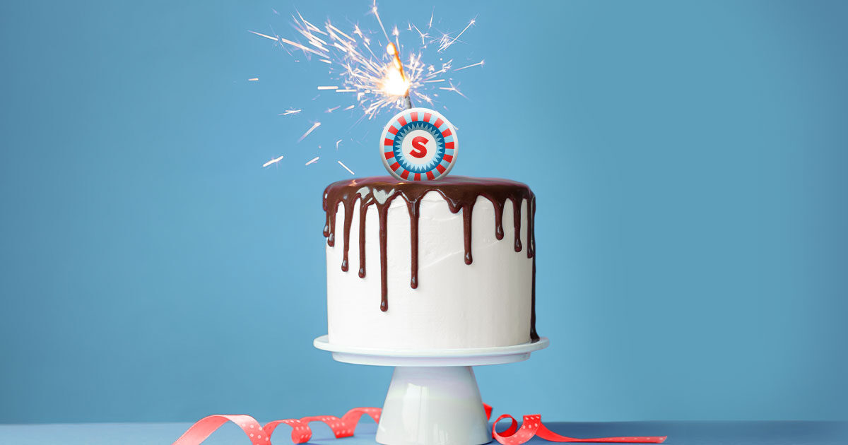 White Birthday cake with chocolate drizzle and a shorelines logo sparkler candle, cake sits on a white cake stand on light blue counter with red ribbon, and light blue background.