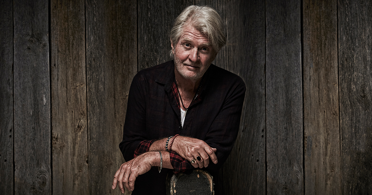 An older man, reminiscent of Tom Cochrane, leaning against a wooden wall.